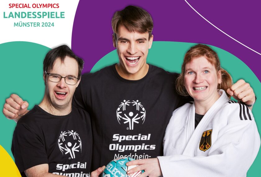 Drei Personen lächeln und posieren für ein Foto. Sie tragen T-Shirts mit dem Special Olympics-Logo und dem Text "Special Olympics Nordrhein-Westfalen". Eine der Personen trägt auch einen weißen Bademantel mit einem deutschen Wappen. Im Hintergrund steht "Special Olympics Landesspiele Münster 2024".