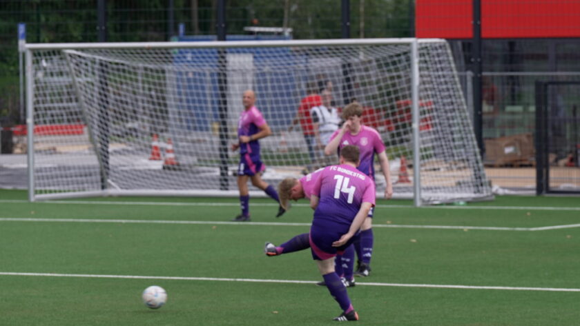 Zwei Fußballspieler in violetten Trikots spielen auf einem grünen Kunstrasenfeld. Ein Spieler mit der Nummer 14 schießt auf das Tor, während ein anderer Spieler in der Nähe steht und beobachtet. Im Hintergrund ist ein Tor mit einem Netz und ein Gebäude zu sehen.