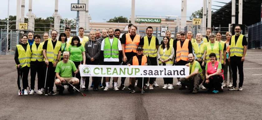 Eine Gruppe von Menschen in gelben und grünen Sicherheitswesten steht vor einem Stadion und hält ein Schild mit der Aufschrift "CLEANUP.saarland". Die Teilnehmer lächeln und posieren für ein Gruppenfoto, während sie sich für eine Säuberungsaktion engagieren.