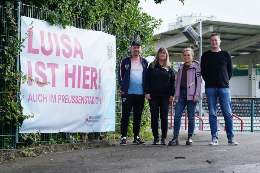 Vier Personen stehen vor einem Zaun im Preußenstadion und lächeln in die Kamera. Hinter ihnen hängt ein großes Plakat mit der Aufschrift "Luisa ist hier! Auch im Preußenstadion" und dem Logo der Beratungsstelle Frauennotruf Münster. Die Personen tragen lässige Kleidung und scheinen sich über das Projekt zu freuen.