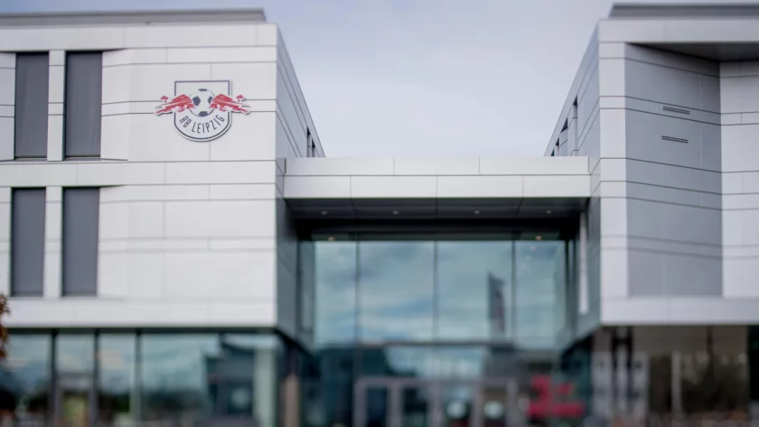 Gebäude mit dem Logo von RB Leipzig, einem Fußballverein, an der Fassade. Das Gebäude hat eine moderne Architektur mit großen Glasfenstern und grauen Paneelen.