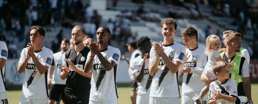 Ein Team von Fußballspielern in weißen Trikots mit schwarzen Akzenten steht auf dem Spielfeld und klatscht. Die Spieler sind in einer Reihe aufgestellt und lächeln. Im Hintergrund sind unscharf die Zuschauer auf den Tribünen zu erkennen.