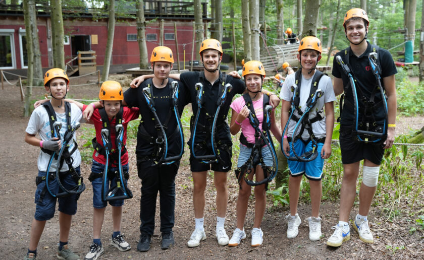 Sechs Kinder stehen in einem Wald und lächeln in die Kamera. Sie tragen Kletterausrüstung, darunter orangefarbene Helme und blaue Klettergurte. Im Hintergrund sind Klettergerüste und ein rotes Gebäude zu sehen.