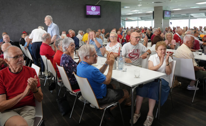 Eine Gruppe älterer Menschen sitzt in einem hellen Raum an Tischen und Stühlen. Sie scheinen an einem geselligen Event teilzunehmen, möglicherweise einem Seniorennachmittag. Viele von ihnen klatschen begeistert, während andere lächeln und sich unterhalten. Einige tragen rote T-Shirts, was auf eine gemeinsame Aktivität oder ein Event hinweisen könnte. Im Hintergrund sind Bildschirme zu sehen, auf denen das Logo der DFL und der Text "1. Bundesliga Seniorenliga" zu erkennen sind.