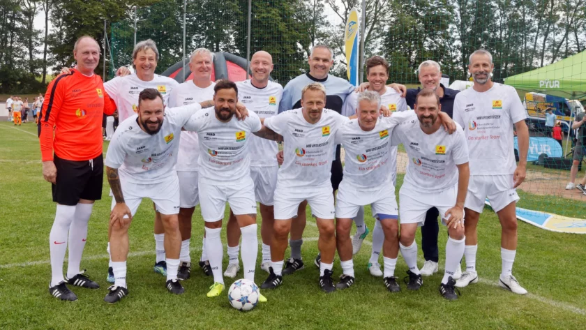 Eine Gruppe von Männern in weißen Trikots und einem Mann in rotem Trikot posieren für ein Gruppenfoto auf einem Fußballfeld. Sie lächeln und haben ihre Arme umeinander gelegt. Die Trikots tragen Aufschriften wie "Wir unterstützen" und "Ein starkes Team". Im Hintergrund sind ein grünes Netz und ein Zelt mit dem Logo "PYUR" zu sehen.