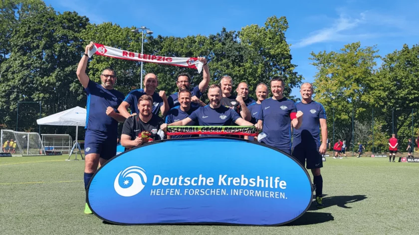 Eine Gruppe von Menschen in blauen Trikots steht vor einem blauen Banner der Deutschen Krebshilfe auf einem Fußballfeld. Sie halten ein Schild mit der Aufschrift "Krebsball Leipzig" und eine rote und weiße Schärpe mit dem Aufdruck "RB Leipzig". Die Gruppe posiert fröhlich und zeigt ihre Unterstützung für die Krebsforschung.