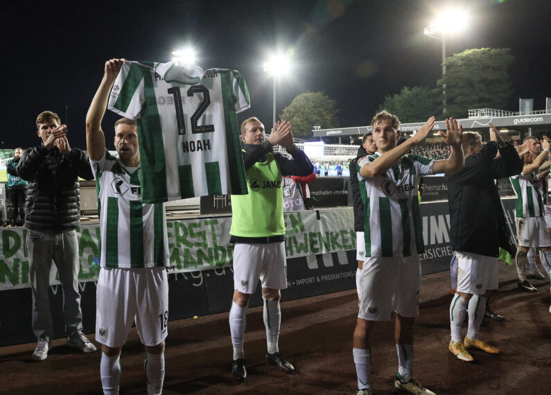 Ein Team von Fußballspielern steht auf dem Spielfeld und hält ein Trikot mit der Nummer 12 und dem Namen "Noah" hoch. Die Spieler tragen grüne und weiße Trikots und sind in einer feierlichen Pose zu sehen, möglicherweise nach einem Sieg oder einem wichtigen Spiel. Im Hintergrund sind die Stadionlichter und einige Zuschauer zu erkennen.