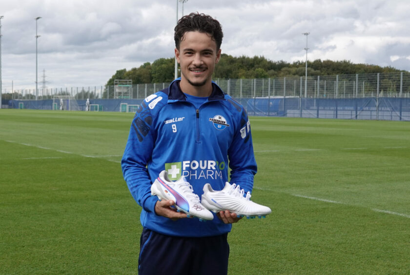 Ein Fußballspieler des SC Paderborn 07 steht auf einem Fußballfeld und hält ein Paar weiße Fußballschuhe in den Händen. Er trägt ein blaues Trainingsoutfit mit dem Logo des Vereins und des Sponsors "Four Pharma". Im Hintergrund sind Tore und eine Umzäunung zu sehen.