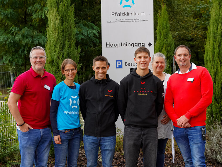 Eine Gruppe von sechs Personen steht vor einem Schild des Pfalzklinikums. Die Personen lächeln und wirken zufrieden. Das Schild weist den Weg zum Haupteingang und zu Besucherparkplätzen. Die Personen tragen verschiedene Kleidungsstücke, darunter T-Shirts und Hoodies mit Logos des Pfalzklinikums und des 1. FC Kaiserslautern.