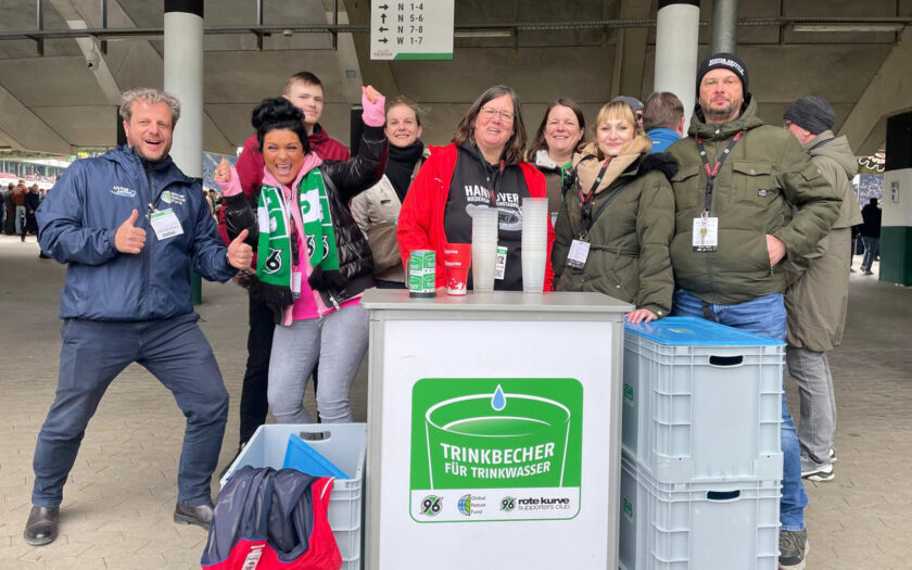 Eine Gruppe von Menschen steht vor einem Stand, der Trinkbecher für Trinkwasser anbietet. Die Personen lächeln und machen Daumen-hoch-Gesten. Der Stand hat ein Schild mit der Aufschrift "Trinkbecher für Trinkwasser" und zeigt Logos von Partnern. Im Hintergrund ist ein Teil eines Stadions zu sehen.