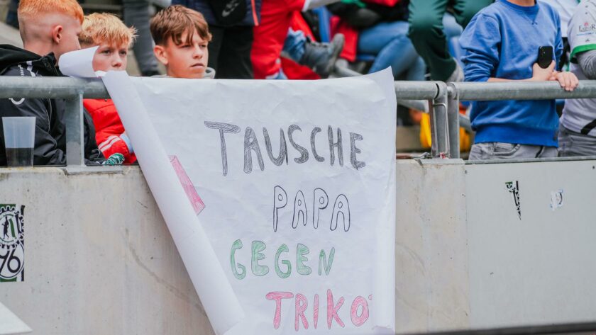 Kinder halten ein selbstgemaltes Schild mit der Aufschrift 'Tausche Papa gegen Trikot' während eines Events, vermutlich dem Weltkinderspieltag, in einem Stadion.