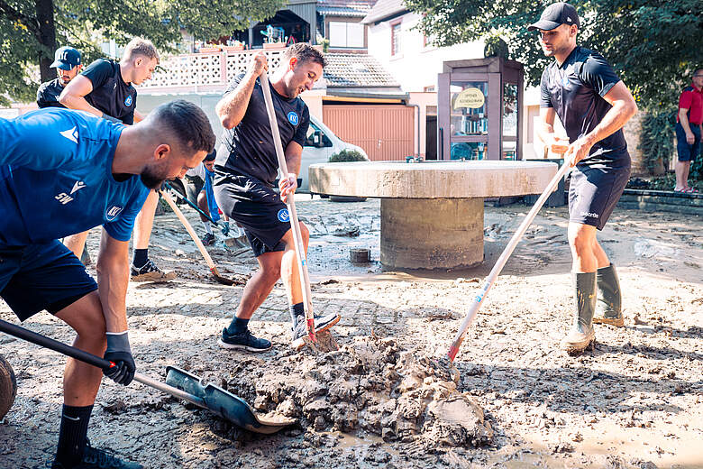 Vier Männer in Sportkleidung arbeiten gemeinsam im Freien, um schlammigen Boden zu bearbeiten. Sie verwenden Schaufeln und andere Werkzeuge, um den Boden zu ebnen und vorzubereiten. Im Hintergrund sind Gebäude und Bäume zu sehen.