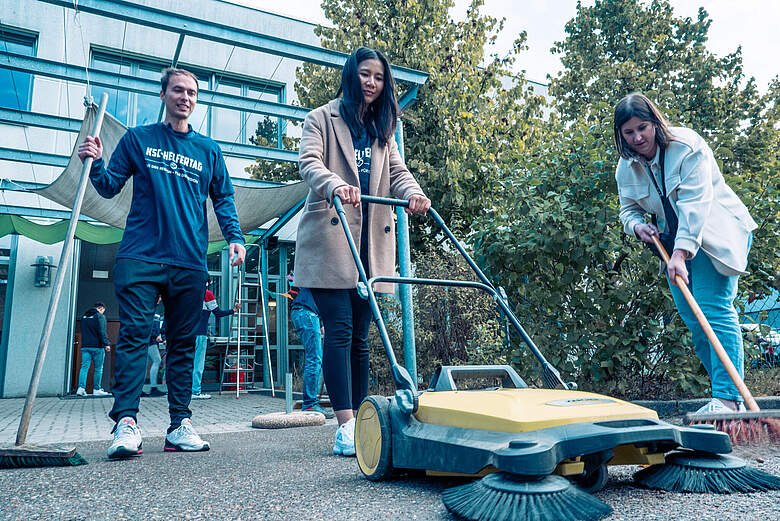 Drei Personen reinigen gemeinsam einen Gehweg mit einem Besen und einem Kehrgerät vor einem Gebäude.