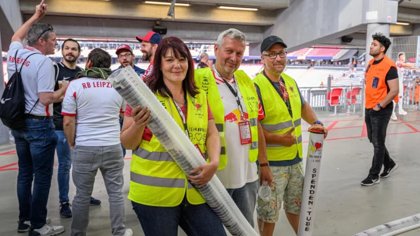Eine Gruppe von Freiwilligen in leuchtend gelben Westen steht in einem Stadion. Sie halten Schläuche und scheinen an einer Spendenaktion teilzunehmen. Die Freiwilligen tragen T-Shirts mit dem Aufdruck "RB Leipzig" und "Team #Brale". Im Hintergrund sind weitere Personen und Teile des Stadions zu sehen.