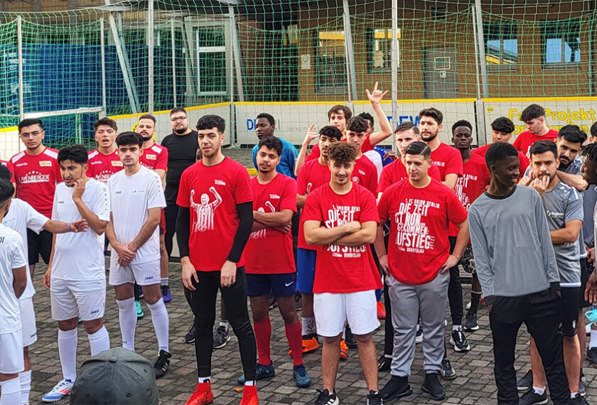 Eine Gruppe von jungen Männern, einige in roten T-Shirts und weiße Sportkleidung, andere in grauen T-Shirts, stehen vor einem Fußballtor. Sie scheinen Teil eines Fußballprojekts zu sein und posieren für ein Gruppenfoto. Im Hintergrund ist ein Gebäude mit einem Schild "Europa Projekt" zu sehen.