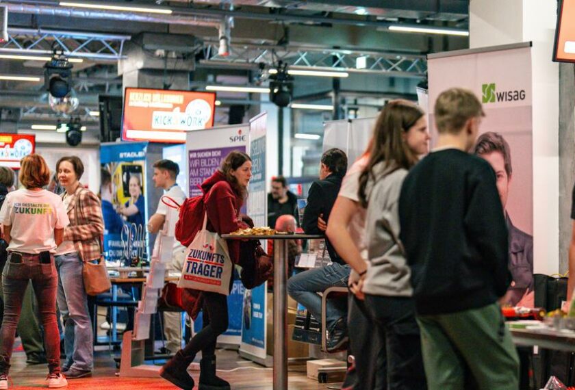 Menschen auf einer Messe oder einem Event, die sich an Ständen informieren und austauschen. Eine Person trägt eine Tüte mit der Aufschrift "Zukunfts Träger". Im Hintergrund sind verschiedene Stände mit Plakaten und Werbematerialien zu sehen, darunter auch ein Stand mit dem Logo von "Kick for Work". Die Atmosphäre wirkt lebhaft und informativ.