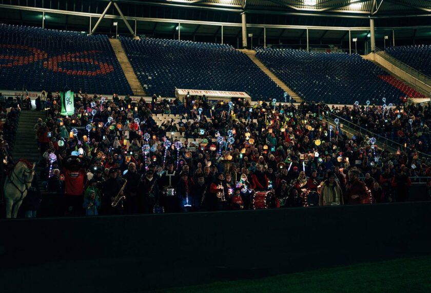 Eine große Menschenmenge nimmt an einem Laternenumzug in einem Fußballstadion teil. Die Zuschauer halten Laternen in verschiedenen Formen und Farben, darunter Sterne, Herzen und Tiere. Die Sitzplätze im Stadion sind größtenteils dunkel, mit Ausnahme einiger beleuchteter Bereiche. Im Vordergrund steht ein Pferd, und eine Person in roter Jacke ist sichtbar. Die Atmosphäre wirkt festlich und gemütlich.