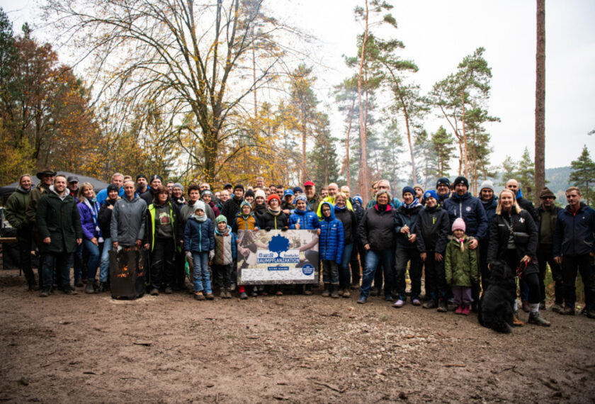 Eine große Gruppe von Menschen steht im Freien und lächelt in die Kamera. Sie halten gemeinsam ein Schild mit der Aufschrift "Baumpflanzaktion" und einem Logo. Die Gruppe besteht aus Erwachsenen und Kindern, die warm gekleidet sind, was auf kühleres Wetter hindeutet. Im Hintergrund sind Bäume und ein bewölkter Himmel zu sehen, was auf einen Wald oder Park als Standort hinweist.