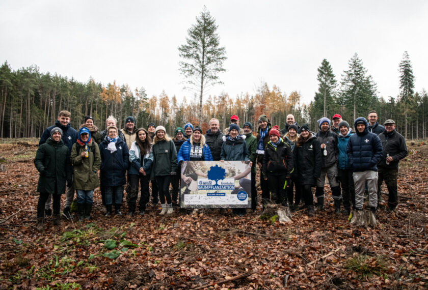 Eine Gruppe von Menschen steht in einem Waldgebiet und nimmt an einer Baumpflanzaktion teil. Sie sind in warme Kleidung gekleidet und lächeln in die Kamera. Im Vordergrund befindet sich ein Schild mit der Aufschrift "Baumpflanzaktion" und verschiedenen Logos. Der Boden ist mit Laub bedeckt und im Hintergrund sind hohe Bäume zu sehen.