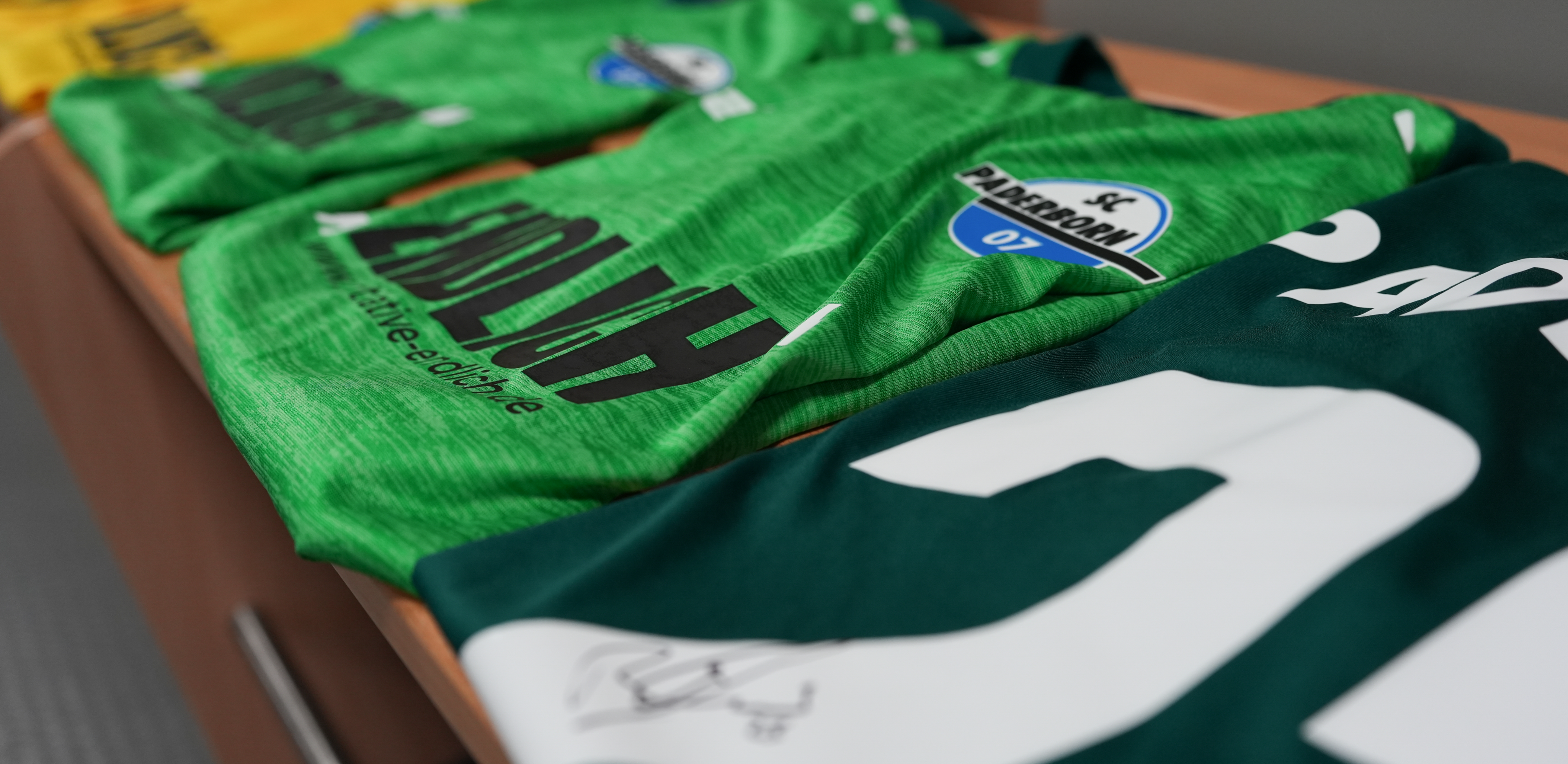 Mehrere grüne Trikots des SC Paderborn liegen auf einem Tisch. Die Trikots sind mit dem Vereinslogo und dem Schriftzug 'Paderborn' bedruckt. Im Hintergrund sind weitere Trikots und ein gelbes Trikot teilweise sichtbar.