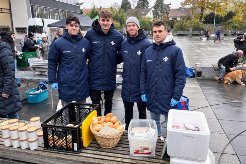 Vier Personen in blauen Jacken stehen vor einem Tisch mit Lebensmitteln. Sie lächeln in die Kamera. Auf dem Tisch befinden sich Becher, Brote, Milch und eine Kühlbox. Im Hintergrund sind weitere Menschen und ein Hund zu sehen.