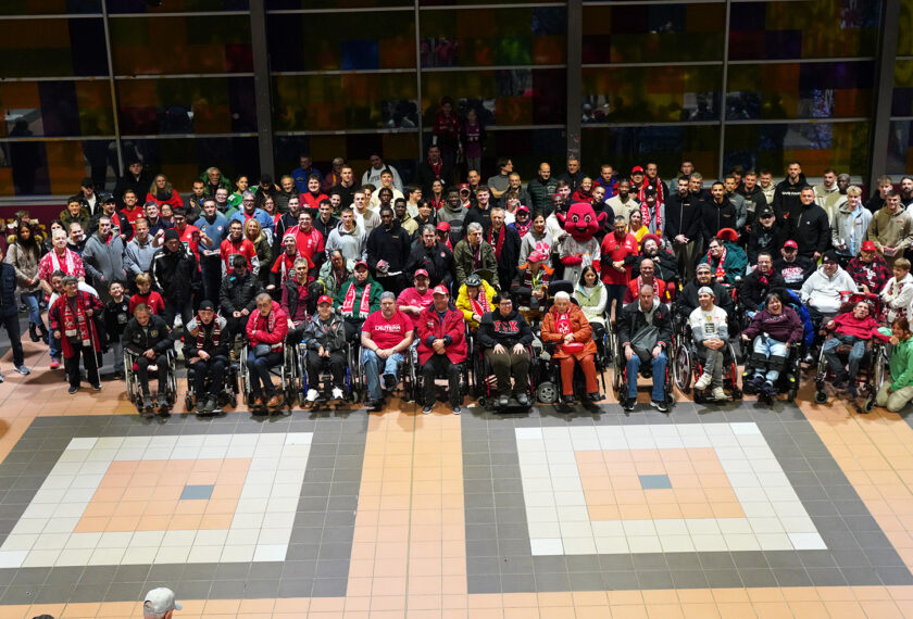 Eine große Gruppe von Menschen, darunter auch Personen im Rollstuhl, steht in einer Halle und posiert für ein Gruppenfoto. Viele tragen rote Kleidung oder Schals, einige halten Schilder oder Maskottchen. Die Atmosphäre wirkt fröhlich und gesellig, möglicherweise bei einem Event oder einer Autogrammstunde des 1. FC Kaiserslautern.