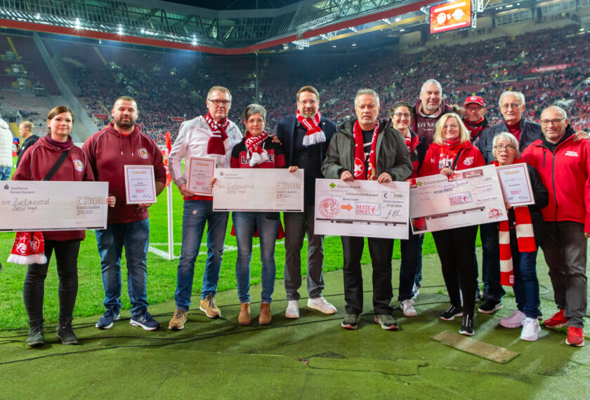Eine Gruppe von Menschen steht auf dem Fußballfeld und hält Schecks in den Händen. Sie tragen verschiedene Kleidungsstücke in den Farben Rot und Schwarz, darunter auch Schals mit Vereinslogos. Im Hintergrund sind Zuschauer auf den Tribünen zu sehen. Die Schecks stammen von der Sparkasse Kaiserslautern und der Glücksbank und sind an verschiedene Empfänger gerichtet, darunter "Betze Engel" und "Betze Ingol". Die Szene findet während eines Fußballspiels des 1. FC Kaiserslautern gegen Paderborn in der 2. Bundesliga, Saison 2024/2025, statt.