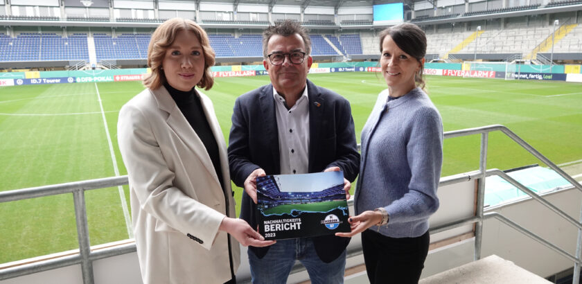 Drei Personen stehen auf einer Tribüne eines Fußballstadions und halten gemeinsam einen Bericht mit dem Titel "Nachhaltigkeitsbericht 2023" des SC Paderborn 07. Im Hintergrund ist der Rasenplatz mit Werbebannern zu sehen.