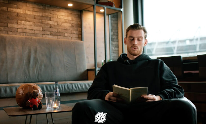 Ein Mann sitzt entspannt in einem modernen Raum und liest ein Buch. Neben ihm auf dem Tisch steht eine Flasche Wasser, eine Kerze und ein Fußball mit einem kleinen Hundespielzeug darauf. Im Hintergrund sind große Fenster mit Blick auf ein Stadion zu sehen.
