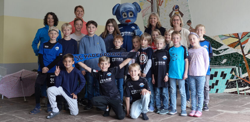 Eine Gruppe von Kindern und Erwachsenen, darunter ein Maskottchen, posieren für ein Gruppenfoto. Sie halten ein Banner mit der Aufschrift "SC Paderborn 07". Die Kinder tragen verschiedene T-Shirts und Hoodies, einige mit Logos von Sponsoren. Die Gruppe steht vor einer bunten Wand mit einem Mosaik.