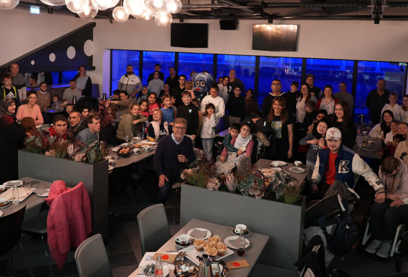 Eine große Gruppe von Menschen sitzt an Tischen in einem Raum mit moderner Beleuchtung und einer großen Fensterfront im Hintergrund. Die Menschen lächeln und scheinen sich zu amüsieren, einige halten Blumensträuße. Der Raum ist mit Tischen und Stühlen ausgestattet, auf denen Snacks und Getränke stehen. Einige Personen sind im Rollstuhl, und alle scheinen Teil einer Feier oder eines besonderen Anlasses zu sein.