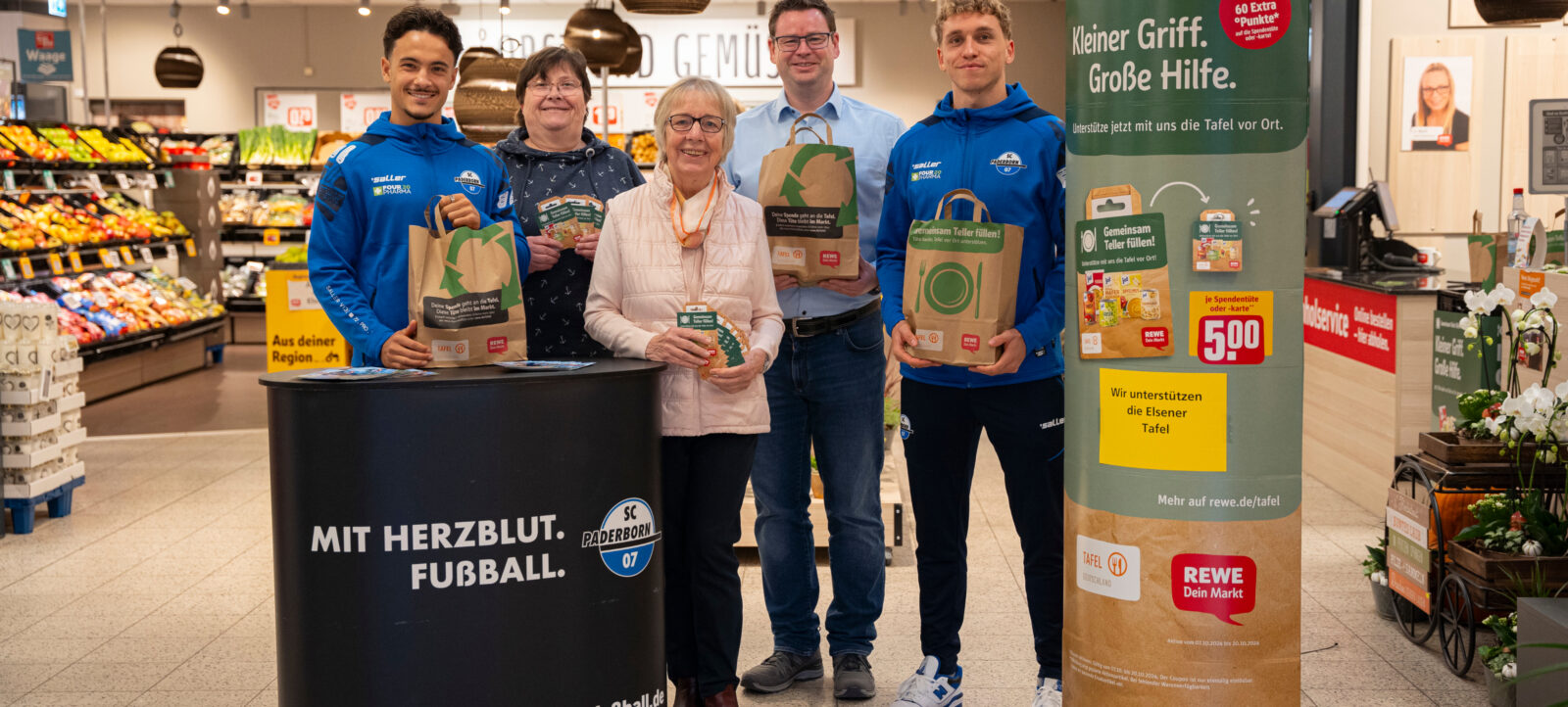 Gruppe von Menschen in einem Supermarkt, die gemeinsam für die Tafel einkaufen. Sie halten Tüten mit Lebensmitteln und stehen vor einem Infostand mit dem Logo des SC Paderborn 07. Der Stand trägt den Slogan 'MIT HERZBLUT. FUSSBALL.' Im Hintergrund sind Regale mit verschiedenen Lebensmitteln zu sehen. Eine Werbesäule informiert über die Aktion 'Kleiner Griff. Große Hilfe.' und die Unterstützung der Elsener Tafel.