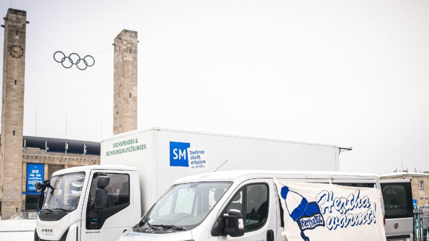 Drei weiße Lieferwagen der Berliner Stadtmission mit dem Logo der Hertha BSC und dem Schriftzug 'Hertha wärmt!' vor dem Olympiastadion Berlin. Im Hintergrund sind die Olympischen Ringe und die Türme des Stadions zu sehen.