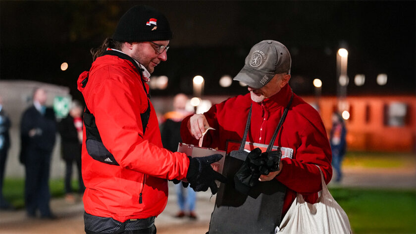 Zwei Personen in roter Kleidung stehen im Freien und tauschen sich aus. Eine Person trägt eine schwarze Mütze mit rotem Logo und eine rote Jacke, während die andere eine graue Baseballkappe und eine rote Jacke trägt. Die Person mit der grauen Kappe hält eine Tüte und einen Gegenstand in den Händen, den die andere Person betrachtet. Im Hintergrund sind weitere Personen und ein beleuchtetes Stadion zu sehen.