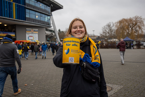 Eine Person steht vor einem Stadion und hält ein gelbes Tuch mit der Aufschrift "Gemeinsam für alle." und einem blauen Logo. Im Hintergrund sind weitere Menschen zu sehen, die sich auf dem Vorplatz des Stadions aufhalten.