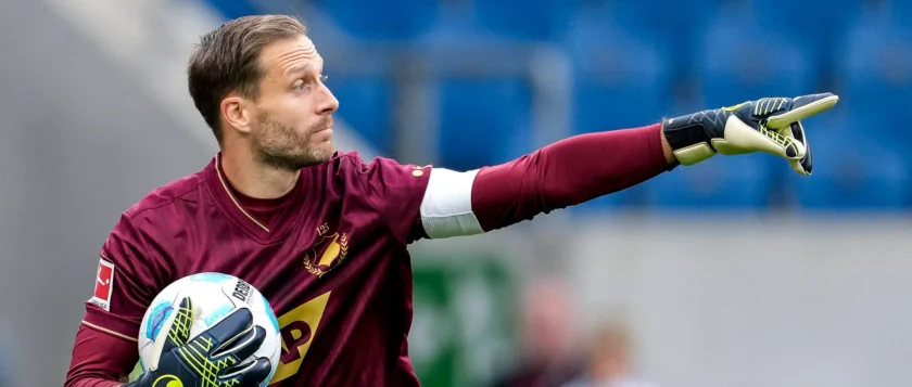 Ein Torwart in maroonfarbenem Trikot und Handschuhen, der einen Ball hält und sich auf dem Spielfeld konzentriert.