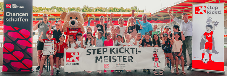 Eine Gruppe von Kindern und Erwachsenen steht vor einem Fußballfeld und hält ein Banner mit der Aufschrift "STEP KICKT!-MEISTER". Ein großes Plüschtier in Form eines Bären mit einem Herz auf der Brust steht in der Mitte der Gruppe. Die Gruppe trägt rote und schwarze Kleidung und hält Urkunden in den Händen. Im Hintergrund sind Tribünen und ein Schild mit der Aufschrift "Chancen schaffen" zu sehen.