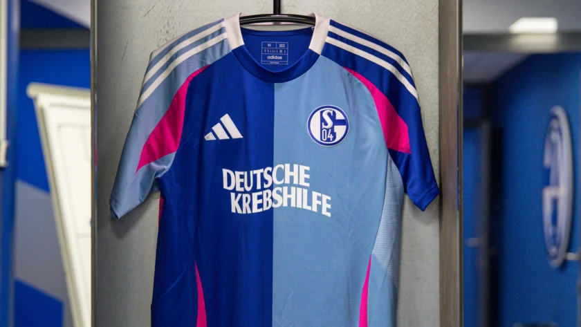 Ein blaues Fußballtrikot mit pinken und weißen Akzenten hängt an einem Haken. Das Trikot hat das Logo des Vereins Schalke 04 und den Schriftzug "Deutsche Krebshilfe" auf der Vorderseite.