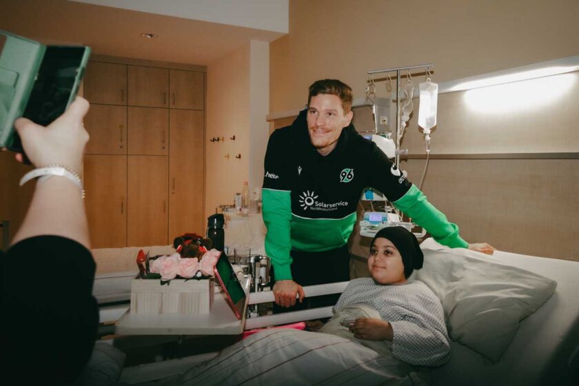 Ein Fußballprofi im Trikot des Vereins mit dem Logo "96" und "SolarService" steht lächelnd an einem Krankenhausbett. Er hält die Hand eines jungen Mädchens, das im Bett liegt und ebenfalls lächelt. Eine Person hält ein Smartphone und macht ein Foto oder ein Video von der Szene. Im Hintergrund sind medizinische Geräte und eine Infusion zu sehen.