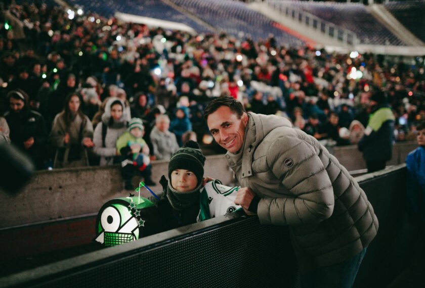Ein Mann und ein Kind sitzen auf einer Stadiontreppe und lächeln in die Kamera. Im Hintergrund ist eine große Menschenmenge zu sehen, die ein Sportevent verfolgt. Das Kind trägt eine Mütze und einen Schal und hält einen kleinen grünen und weißen Fan-Artikel. Der Mann trägt eine graue Daunenjacke.