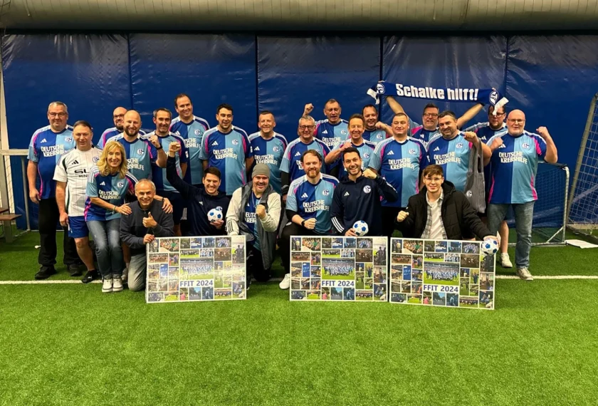 Eine Gruppe von Menschen in Schalke-Trikots steht vor einem Tor im Fußballstadion. Sie halten Plakate mit Fotos von Fußballspielen und dem Text "FFIT 2024". Die Atmosphäre ist fröhlich und gesellig, mit einigen Personen, die Daumen hoch zeigen.