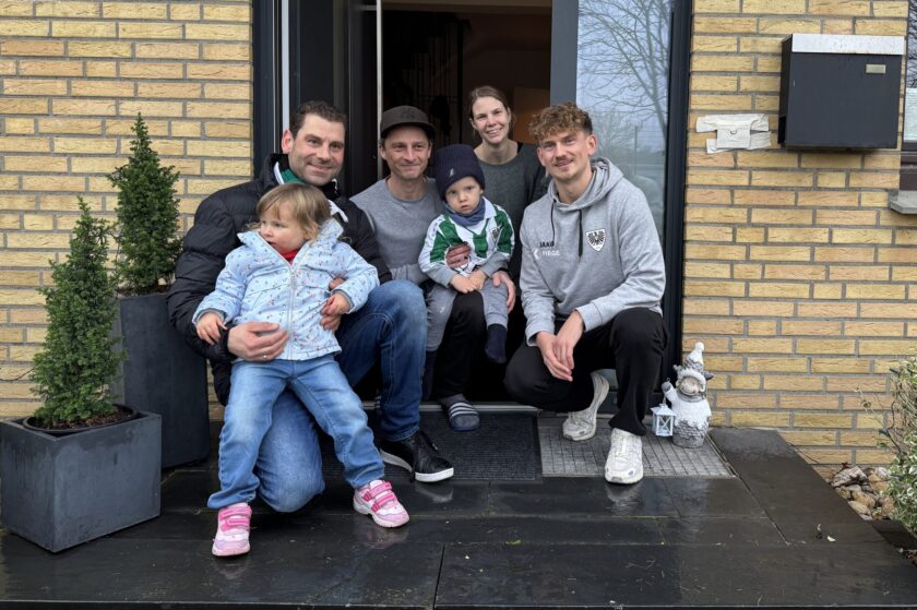 Eine Familie mit fünf Personen und einem Fußballspieler posieren vor einem Hauseingang. Die Familie besteht aus zwei Erwachsenen und drei Kindern, die auf den Stufen vor der Tür sitzen. Der Fußballspieler, der einen Hoodie mit einem Vereinslogo trägt, sitzt neben den Kindern. Alle lächeln und wirken fröhlich. Im Hintergrund ist eine gelbe Backsteinwand mit einem Briefkasten und einigen Pflanzen in Töpfen zu sehen.