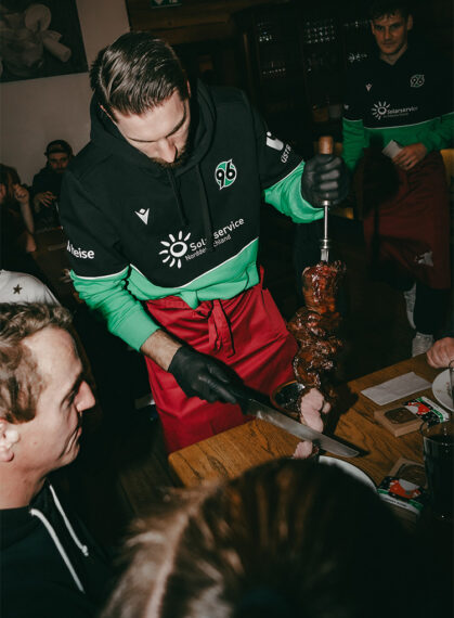 Ein Mann in einer schwarzen Jacke mit grünen und roten Ärmeln und einem roten Schürze schneidet Fleisch von einem Spieß. Er trägt Handschuhe und hält ein Messer in der Hand. Im Hintergrund sind weitere Personen zu sehen, die ebenfalls ähnliche Kleidung tragen. Die Szene findet in einem Raum mit Holztischen und Stühlen statt, und es scheint sich um eine Veranstaltung oder ein Event zu handeln.