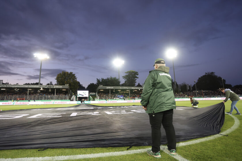 Drei Personen bereiten ein Fußballfeld vor, indem sie eine schwarze Plane auf den Rasen legen. Die Szene spielt bei Dämmerung oder in der Nacht, da die Flutlichtmasten eingeschaltet sind und das Spielfeld beleuchten. Im Hintergrund sind Zuschauer auf den Tribünen zu sehen, die das Geschehen verfolgen. Die Personen tragen grüne Jacken, auf denen das Wort "Sadlmeier" zu erkennen ist.