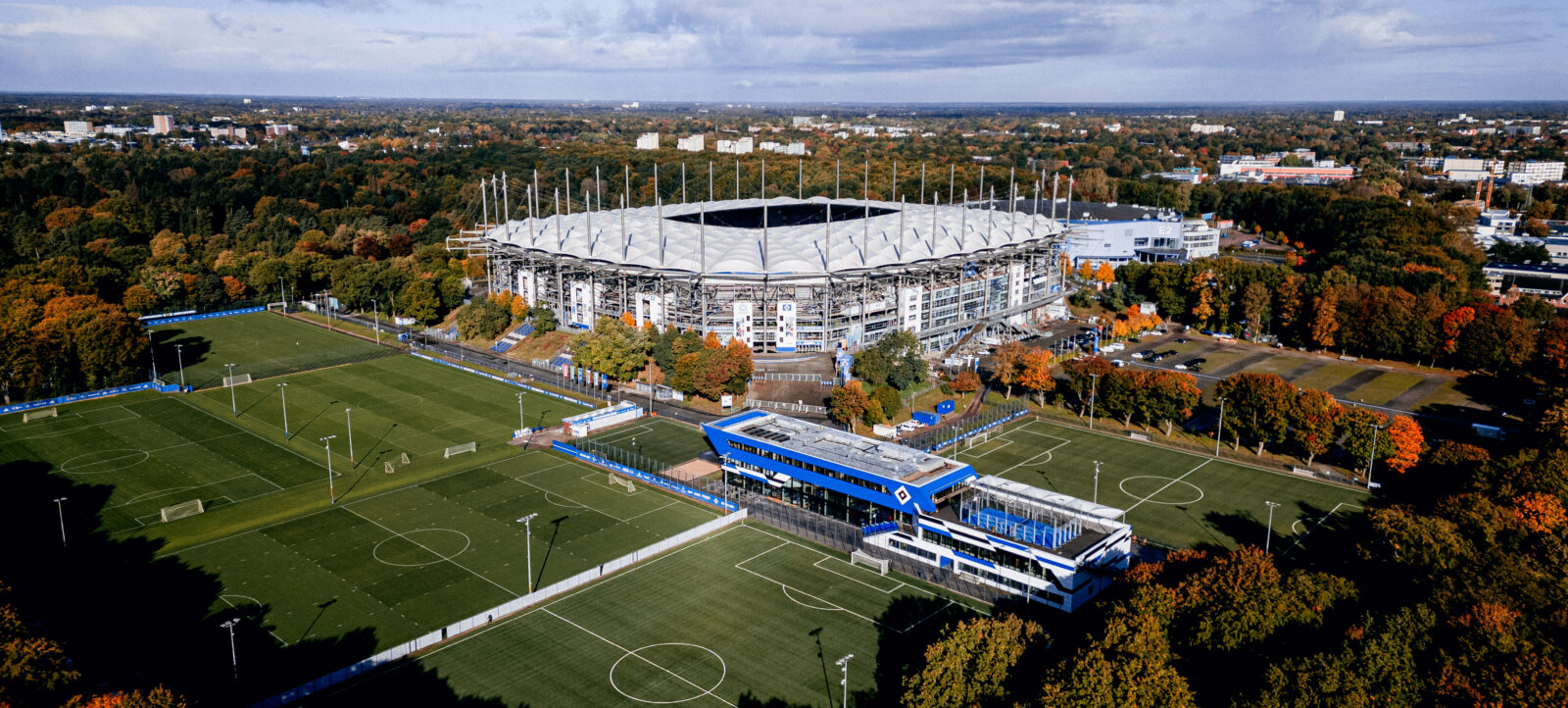 Luftaufnahme des Volksparkstadions in Hamburg, Deutschland, am 11.10.2024. Das Bild zeigt das große Stadion mit seinem charakteristischen Dach und den umliegenden Bereichen, darunter Trainingsplätze und ein kleineres Stadion. Die Herbstfarben der umliegenden Bäume sind deutlich sichtbar, und der Himmel ist teilweise bewölkt.
