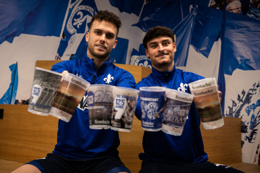 Zwei Fußballspieler in blauen Trikots mit dem Logo des 1. FC Köln halten jeweils zwei Krügerl mit dem Logo des Vereins und der Aufschrift "Krombacher". Sie sitzen vor einem Hintergrund mit blau-weißen Fahnen und dem Vereinswappen.