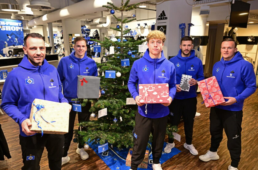 Vier Männer in blauen Hoodies stehen vor einem geschmückten Weihnachtsbaum in einem Fußball-Fanshop. Sie halten jeweils ein Geschenk in den Händen und lächeln in die Kamera. Die Männer sind von links nach rechts: Torwarttrainer Sven Hoeh, Torwart Matheo Raab, Torwart Hannes Hermann, Torwart Daniel Heuer Fernandes und Torwart Tom Mickel. Der Weihnachtsbaum ist mit blauen und weißen Dekorationen geschmückt, darunter auch kleine Fußball-Elemente. Im Hintergrund sind Regale mit Fußball-Artikeln und ein Schild mit der Aufschrift "HSV Shop" zu sehen.