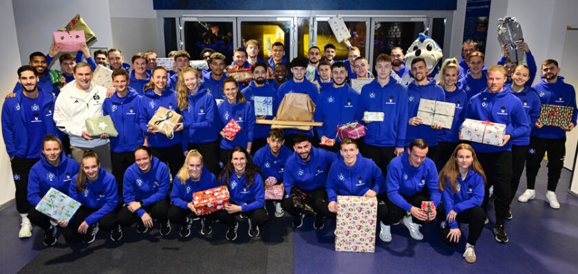 Eine große Gruppe von Menschen in blauen Team-Jacken steht für ein Gruppenfoto. Sie halten verschiedene Geschenke und Päckchen in den Händen und lächeln in die Kamera. Im Hintergrund sind weitere Personen und ein Maskottchen zu sehen. Die Atmosphäre wirkt fröhlich und festlich, vermutlich im Rahmen einer Weihnachtsfeier.