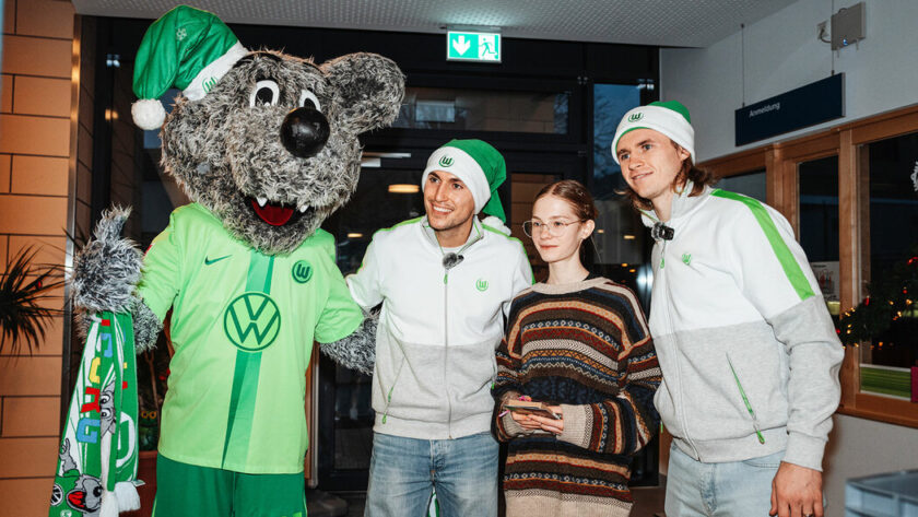 Vier Personen, darunter ein Maskottchen im grünen Trikot mit Volkswagen-Logo, stehen in einem Flur und lächeln in die Kamera. Alle tragen Weihnachtsmützen.
