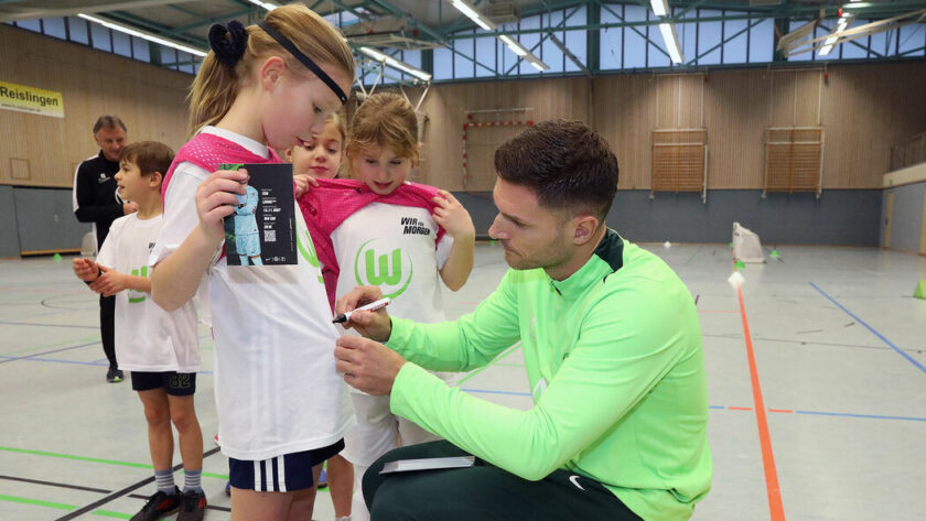 Ein Trainer in grüner Sportkleidung interagiert mit drei Kindern in einer Sporthalle. Die Kinder tragen weiße T-Shirts mit einem grünen Logo und halten ein Tablet. Der Trainer zeigt auf das Tablet, während die Kinder aufmerksam zuschauen. Im Hintergrund sind weitere Kinder und ein Erwachsener zu sehen.